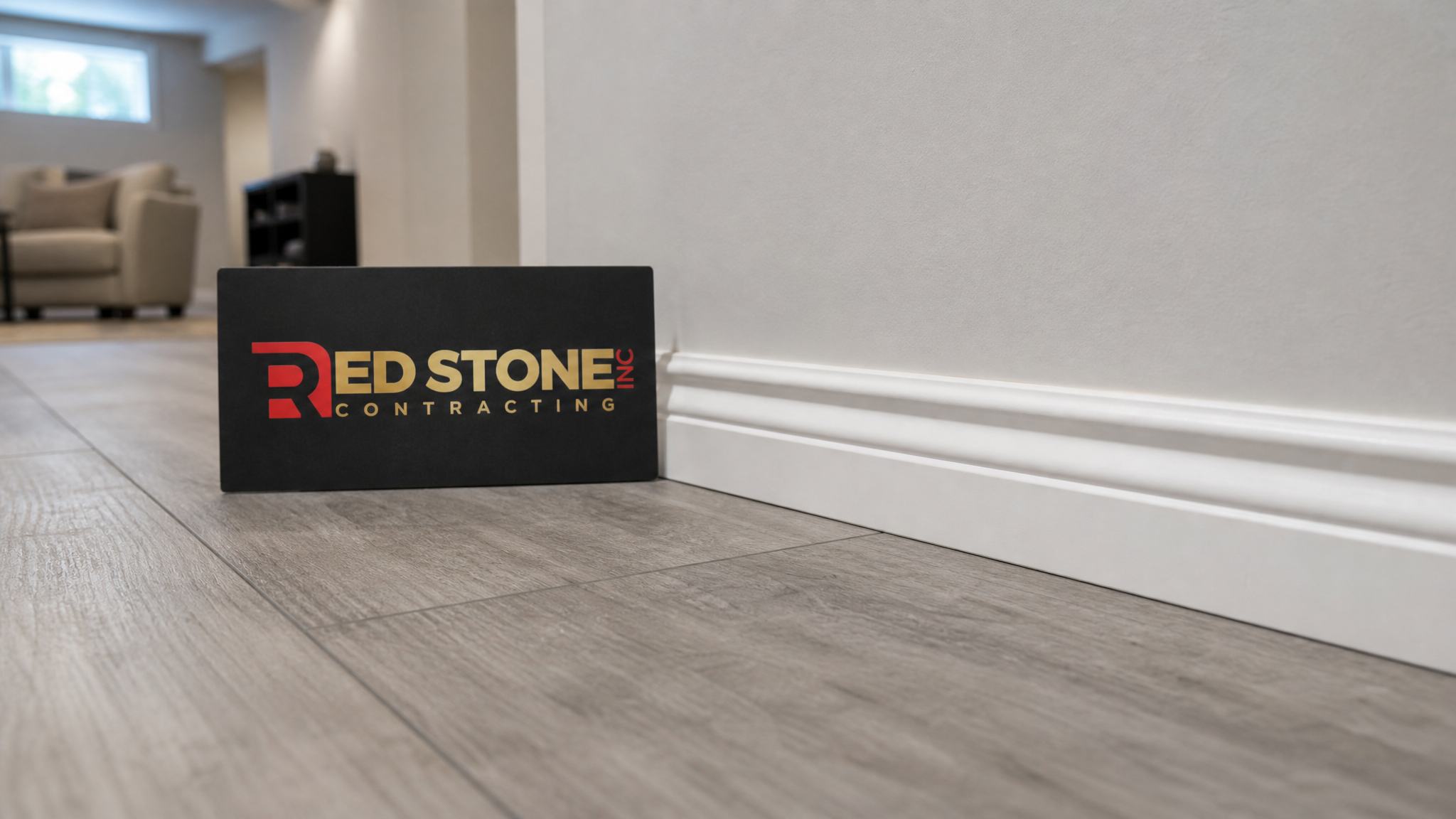 Close-up of luxury vinyl plank flooring and clean trim work in a finished Burlington basement with a Red Stone Contracting placard