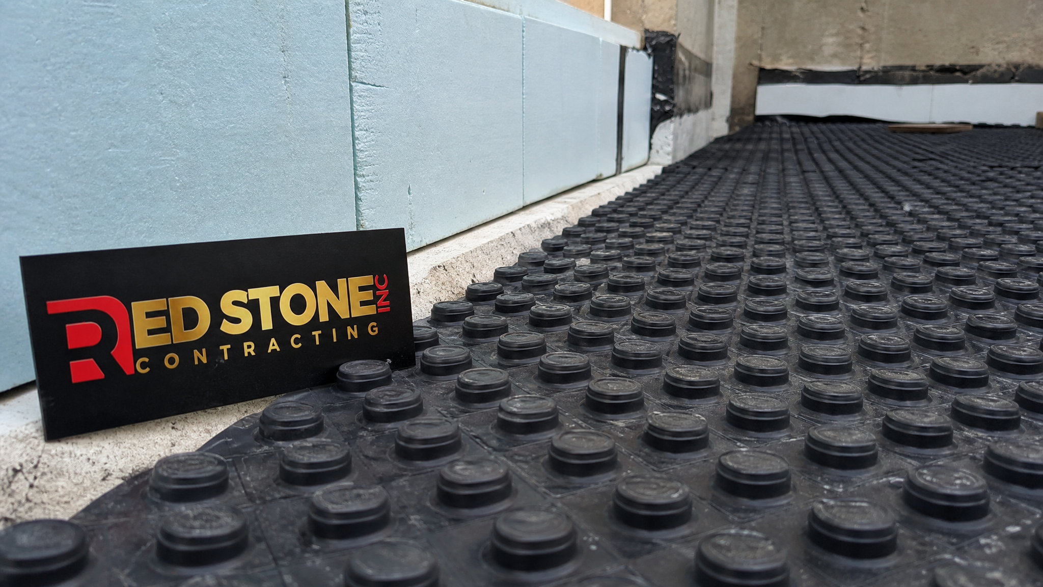 Close-up of a dimpled subfloor membrane and below-grade insulation with a small Red Stone Contracting sign placard nearby